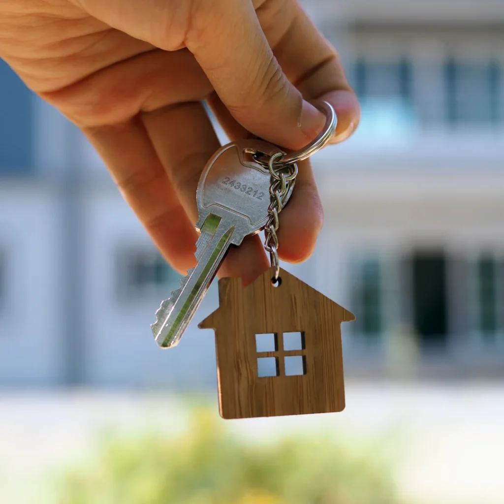 House key on a wooden house-shaped keychain held in front of a home