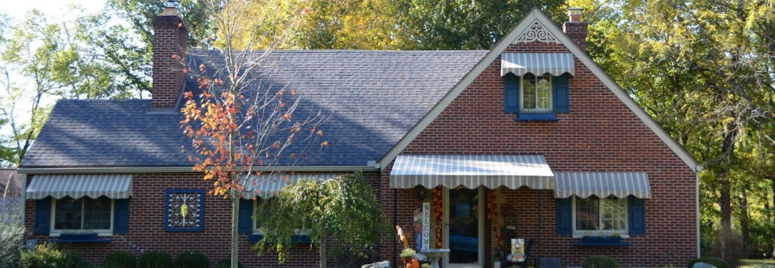 Red brick Cape house with blue shutters and striped awnings in early fall, the kind of home sold in Albany, NY