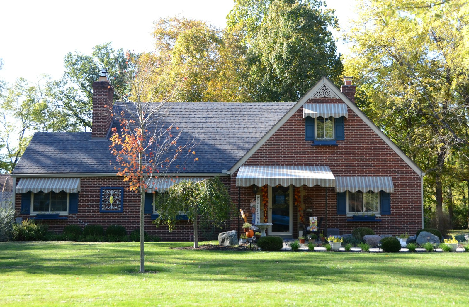 Red brick Cape house with blue shutters and striped awnings in early fall, the kind of home sold in Albany, NY