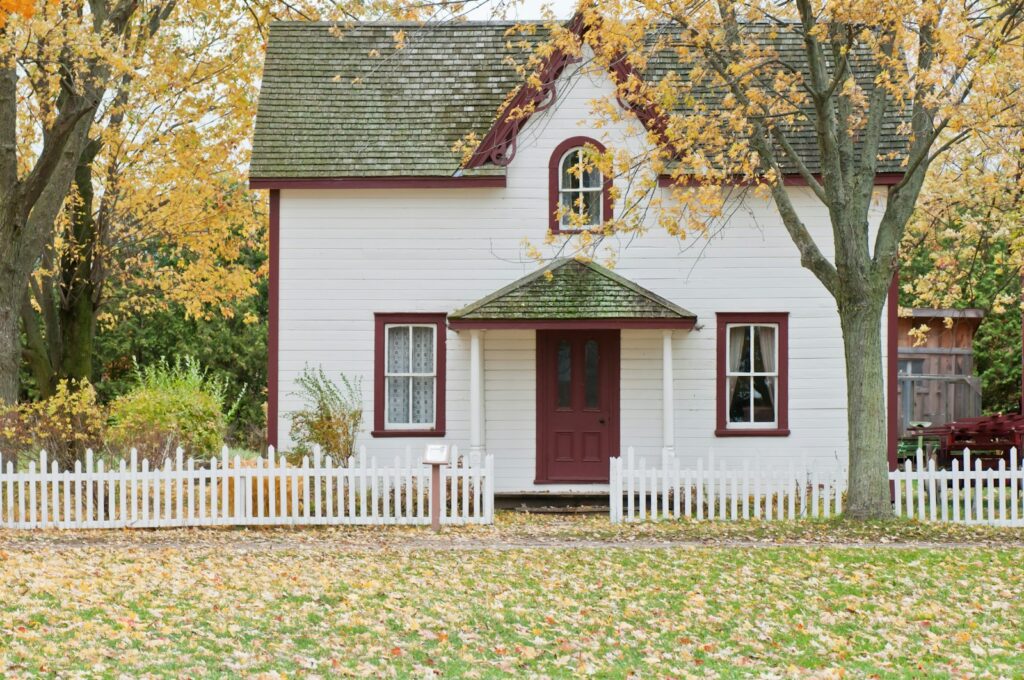 White clapboard house with red trim and a picket fence under yellow autumn maples