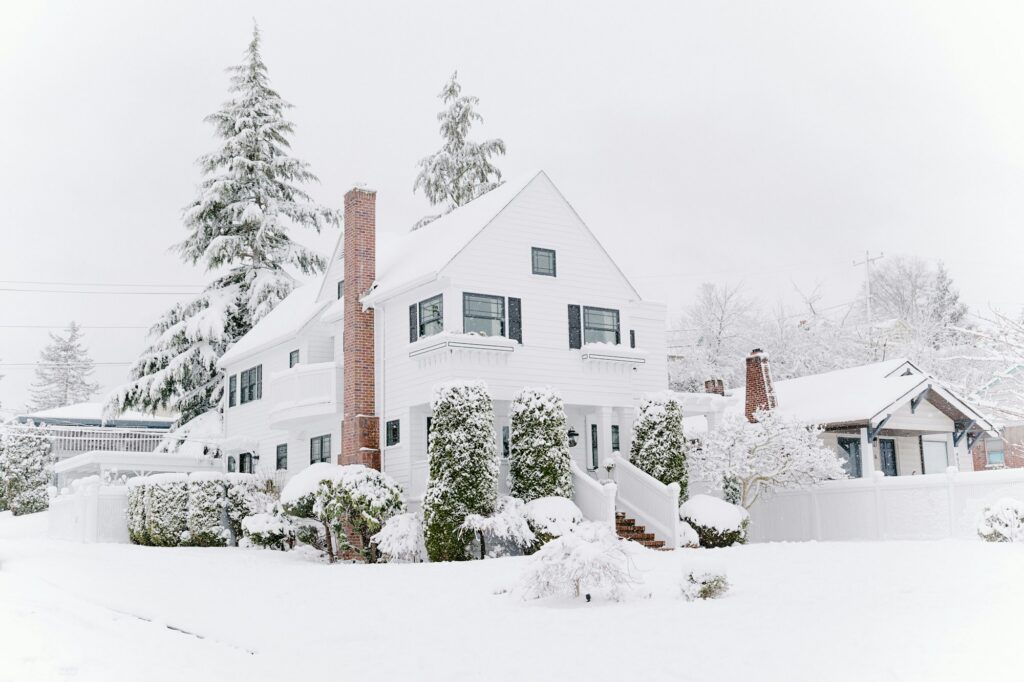 White two-story colonial house with a brick chimney and snow-covered yard and evergreens