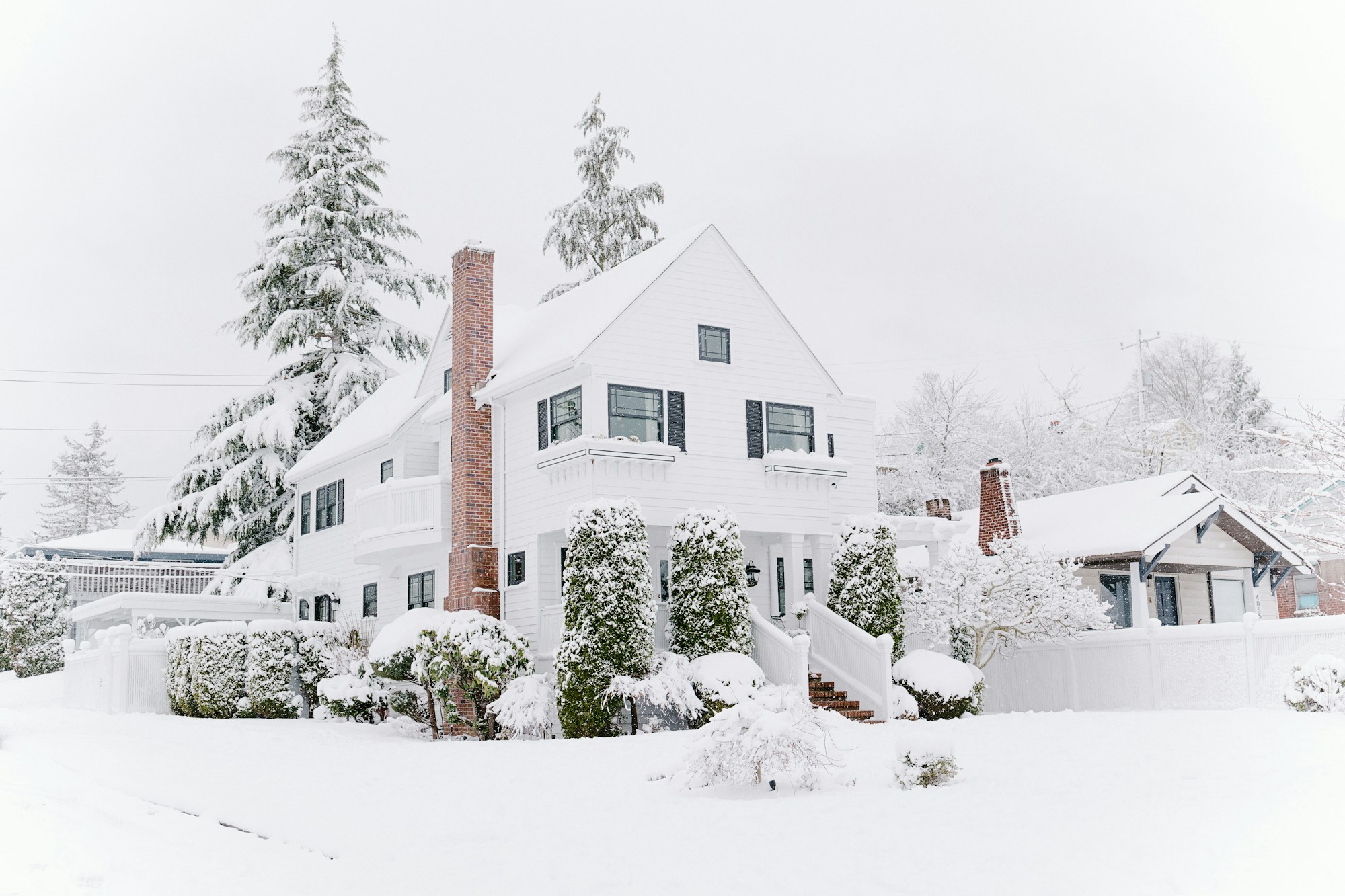 White two-story colonial house with a brick chimney and snow-covered yard and evergreens